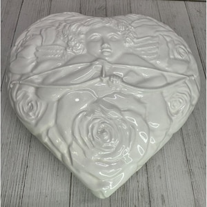 Puede incluir: Plato de cerámica blanco en forma de corazón con un diseño en relieve. El diseño presenta un querubín sosteniendo un arco y una flecha, rodeado de rosas. El plato tiene un acabado brillante.