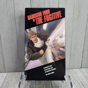 May include: A VHS tape case for the movie "The Fugitive" starring Harrison Ford. The cover features a photo of Harrison Ford running through a train car. The text on the case reads "A murdered wife. A one-armed man. An obsessed detective. The chase begins."