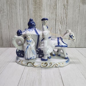 May include: A porcelain figurine of a horse-drawn carriage with two figures in blue and white. The carriage is decorated with blue and white floral designs. The figures are dressed in blue and white clothing. The figurine is on a white base with gold accents.