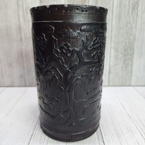 May include: A dark brown cylindrical wooden brush pot featuring intricate relief carvings of a landscape scene. The surface displays detailed depictions of mountains, stylized trees, and clouds, with a smooth, circular rim at the top edge.