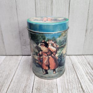 Puede incluir: Una lata vintage con tapa azul y una ilustración colorida de Santa Claus llevando un saco de regalos y caminando por un bosque nevado.