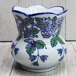 May include: A white ceramic vase with a scalloped rim and a hand-painted floral pattern featuring purple flowers, yellow centers, and green leaves. The vase has a rounded body and a blue painted trim along the top edge and the base.