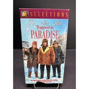 Puede incluir: Una cinta VHS de la película Trapped in Paradise protagonizada por Nicolas Cage, Jon Lovitz y Dana Carvey. La portada muestra a los tres actores con ropa de invierno en un entorno nevado, con el logo 20th Century Fox Selections en la parte superior.