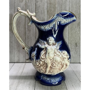 May include: A vintage ceramic pitcher with a dark blue glaze. The pitcher features a cream-colored handle and relief details of cherubs and floral patterns. The pitcher is approximately 8 inches tall.