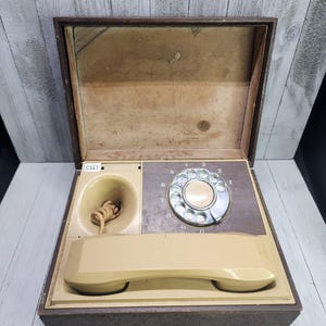 May include: A vintage rotary dial phone in a brown wooden case. The phone has a beige receiver and a beige rotary dial with black numbers. The case has a brown felt lining.
