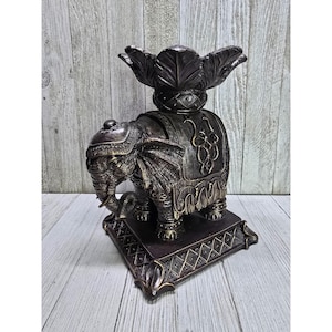May include: A bronze-colored elephant figurine with intricate detailing. The elephant has a decorative saddle and a floral-shaped top. The base is square with a diamond pattern. The figurine is made of a metallic material.