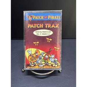 Op de afbeelding: Een doorzichtig plastic cassettehoesje met de albumhoes van Patch the Pirate Patch Trax Vol. 17 Issues 1-3. De illustratie toont een cartoon tuba, viool en fluit met muzieknoten tegen een rode en gele achtergrond van een podium.
