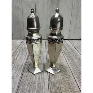 Peut inclure: Une paire de salières et poivrières argentées vintage. Chaque shaker a un dessus conique avec des trous, une bande décorative et une base à facettes. Les shakers sont présentés sur une surface en bois.