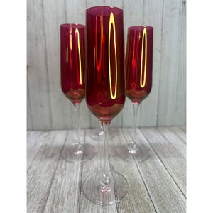 May include: Four ruby red champagne flutes with clear stems and bases. The glasses have a tall, slender design and a metallic gold rim. The flutes are arranged on a light wood-grain surface.