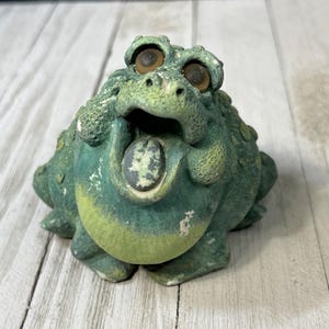 May include: A green ceramic frog figurine with large brown eyes and a wide open mouth. The frog has a textured surface and a green tongue sticking out.