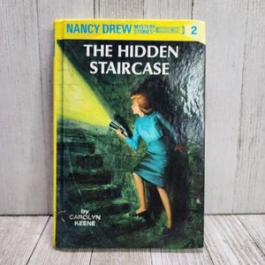 Könnte beinhalten: Ein gelbes und schwarzes Taschenbuch mit dem Titel "The Hidden Staircase" von Carolyn Keene. Das Buch ist Teil der Nancy Drew Mystery Stories-Reihe und Nummer 2 der Reihe. Das Cover zeigt eine Illustration von Nancy Drew, die eine Taschenlampe hält und eine dunkle Treppe hinuntergeht.