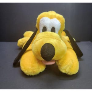 May include: A plush yellow dog toy with a black nose, black ears, and a pink tongue. The dog has white around its eyes and is lying down. This soft toy is a classic character.