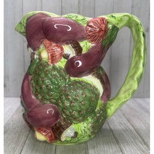 May include: A ceramic pitcher with a green handle and a body decorated with colorful vegetables. The pitcher features artichokes, eggplants, and mushrooms in shades of green, purple, and brown. The pitcher is a decorative kitchenware item.