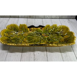 May include: A long, rectangular ceramic serving dish with a leaf design. The dish is primarily green and yellow with a black accent. The leaf pattern is detailed with visible veins, and the edges are scalloped.