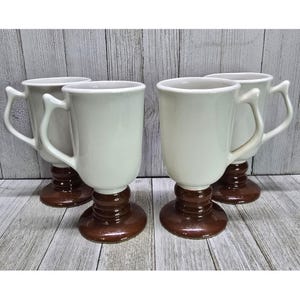 Może przedstawiać: Cztery białe ceramiczne kubki do kawy z brązowymi podstawami i dużymi uchwytami. Kubki mają klasyczny design, odpowiedni do gorących napojów. Kubki są prezentowane na jasnej drewnianej powierzchni.