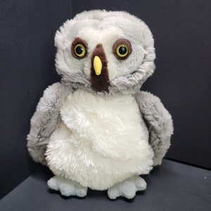 May include: A gray and white plush owl with brown eyes and a yellow beak. The owl is sitting on a black surface.