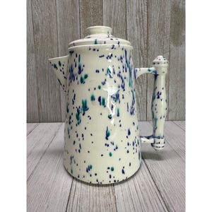Op de afbeelding: Crèmekleurige keramische koffiepot met een deksel, tuit en handvat. De pot is versierd met blauwe en teal spetters. Het handvat is aan de zijkant van de pot bevestigd. De pot staat op een houten oppervlak.