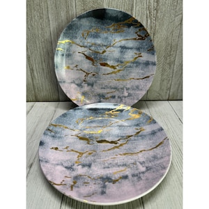 May include: Two round plates featuring a marbled pattern in shades of dusty pink, slate gray, and deep charcoal. The surface of each plate is decorated with thin, irregular metallic gold veins that mimic the appearance of natural stone veining.