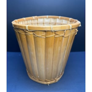 May include: A cylindrical wastebasket constructed from vertical bamboo slats with a light tan finish. The structure features a reinforced circular rim and base, with thin, flexible bamboo strips woven horizontally to secure the vertical slats in place.