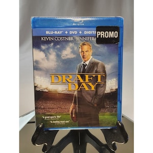 Puede incluir: Una caja de disco Blu-ray con una pegatina promocional. La caja presenta una foto de Kevin Costner y el título "Draft Day".
