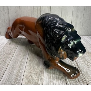 Puede incluir: Figura de león de cerámica en posición reclinada. El león tiene un cuerpo marrón brillante y una melena negra. La figura está colocada sobre una superficie de madera.