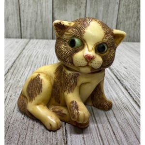 May include: A vintage figurine of a seated cat with a cream-colored body and brown markings. The cat has large green eyes, a pink nose, and a detailed fur pattern. The figurine is made of a glossy material and is posed in a sitting position.