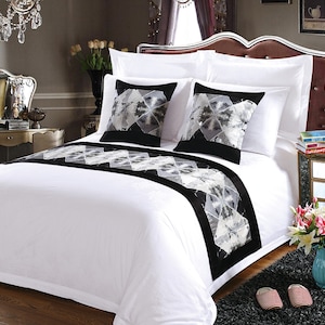 May include: White bedding with a black and white patterned bed runner. The bed has two white pillows and two black and white patterned pillows. The bed is in a bedroom with a nightstand and a lamp.