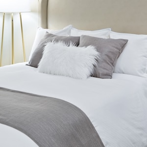 May include: A white bed with a gray bed runner and a white fluffy pillow on top of a gray pillow. The bed is made with white sheets and a white duvet cover.