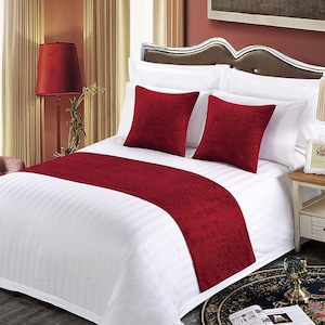 May include: A white bed with a red velvet bed runner and two red velvet throw pillows. The bed is made with crisp white sheets and a white duvet cover. The bed is in a bedroom with a nightstand and a lamp.