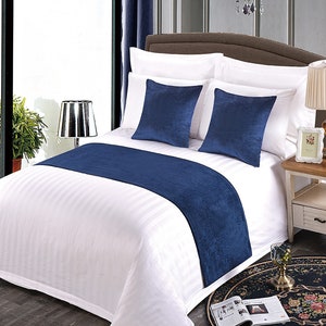 May include: A bed with white striped bedding, two navy blue square pillows, and a navy blue bed runner. A small side table with a lamp and a framed picture is next to the bed. The room has blue curtains.