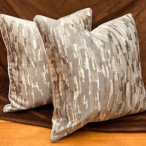 May include: Two decorative throw pillows with a gray, brown, and white abstract pattern. The pillows have a white piping around the edges.