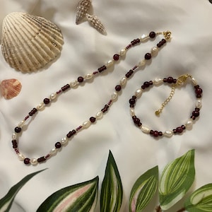 Könnte beinhalten: Ein Halsketten- und Armband-Set mit weißen Perlen, dunkelroten Perlen und goldfarbenen Verschlüssen. Der Schmuck wird auf einer weißen Oberfläche mit Muscheln und Grünzeug im Hintergrund präsentiert. Die Perlen sind klein und rund.