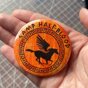 May include: A round orange and black button with the text "Camp Half-Blood" and a black silhouette of a winged horse with a Greek key border.