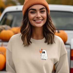 Könnte beinhalten: Beiges Sweatshirt mit einer kleinen Geistergrafik, die ein kürbisförmiges Getränk hält. Der Geist ist weiß mit schwarzen Details. Der Hintergrund umfasst Kürbisse und einen weißen LKW, was auf ein Herbst- oder Halloween-Thema hindeutet.