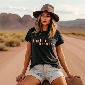Könnte beinhalten: Eine Frau trägt ein schwarzes T-Shirt mit dem Text "tutto bene" in pfirsichfarbenen Buchstaben. Sie trägt einen braunen Fedora-Hut und eine hellgraue Jeans-Shorts. Sie sitzt auf einem Felsen in einer Wüstenlandschaft.