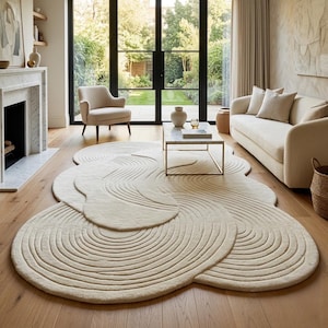 May include: An off-white area rug with an irregular, organic shape featuring a textured pattern of concentric, raised circular lines that create a ripple effect across the surface, covering a large portion of the wooden floor in the room.