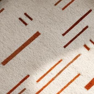 May include: Close-up of a beige rug with a repeating pattern of brown and orange diagonal lines. The lines vary in length and thickness, creating a geometric design. The rug has a soft, textured appearance.