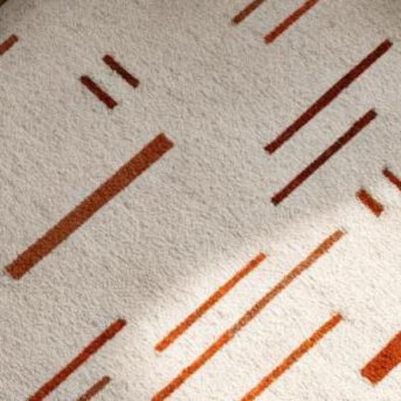 May include: Close-up of a beige rug with a repeating pattern of brown and orange diagonal lines. The lines vary in length and thickness, creating a geometric design. The rug has a soft, textured appearance.