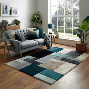 May include: A contemporary living room scene with a grey sofa, patterned cushions, and a geometric rug in blue, grey, and beige. The room has a large window and wooden flooring.
