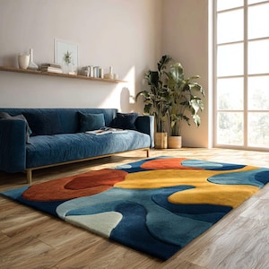 May include: A colourful, abstract rug with a pattern of overlapping shapes in blue, yellow, and rust. The rug is placed on a wooden floor in a living room, with a blue sofa and large windows visible. The rug's design adds a modern touch to the room.