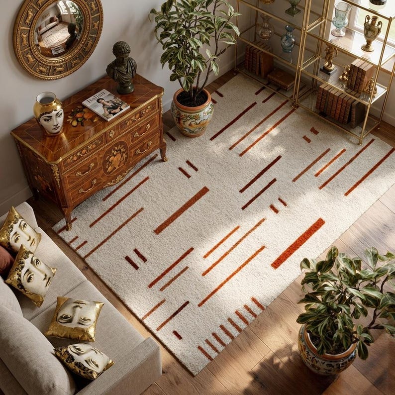 May include: An overhead shot of a living room with a cream-colored rug featuring a geometric pattern of brown lines. The room includes a wooden dresser, a sofa with decorative pillows, and potted plants. A gold-framed mirror and a bookshelf are also visible.