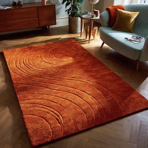 Peut inclure: Un tapis orange brûlé avec un motif tourbillonnant texturé. Le tapis rectangulaire est fait d'un matériau doux et présente un motif de lignes courbes. Le tapis est posé sur un sol en bois, ajoutant de la chaleur à la pièce.