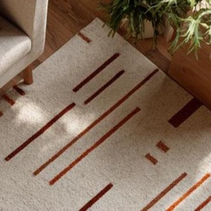 May include: Beige rectangular rug with a geometric pattern of rust-colored lines. The rug is placed on a wooden floor, with a glimpse of a chair and potted plants in the background. The rug's design adds a modern touch to the room.
