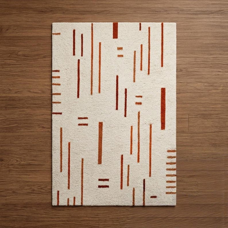 May include: Rectangular off-white rug with a repeating pattern of vertical and horizontal rust-colored lines. The rug is placed on a wooden floor. The design is modern and geometric, suitable for a living room or bedroom.