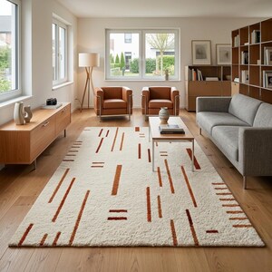 May include: A cream-colored area rug with a geometric pattern of rust-colored lines and dashes. The rug is placed in a modern living room with hardwood floors, a gray sofa, and two brown leather armchairs.
