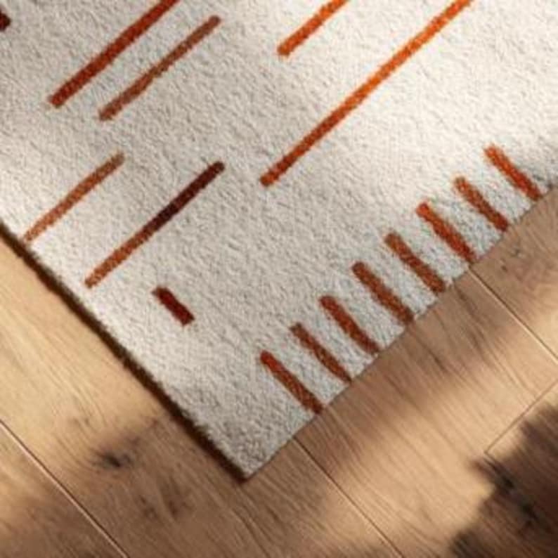 May include: Close-up of a beige rug with a geometric pattern of rust-colored lines. The rug is placed on a light-colored wooden floor. The rug's texture is visible, and the lines are evenly spaced.