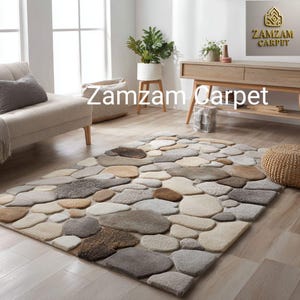 May include: A rectangular area rug featuring a textured, multi-dimensional pattern of rounded, pebble-like shapes in various shades of gray, beige, cream, and brown. The text Zamzam Carpet is visible in the upper right corner of the image.