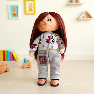 May include: A handmade crochet doll with long auburn hair, wearing a gray sweatshirt and pants set with a Mickey Mouse pattern. The doll has a matching gray purse with a Mickey Mouse design and is wearing black shoes.