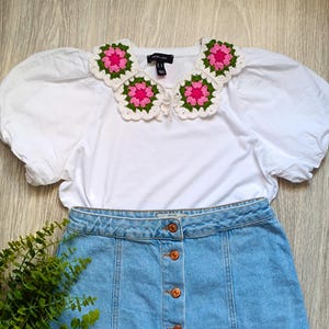 May include: A white blouse with puffy sleeves and a crocheted collar featuring pink and green floral designs. Paired with a light blue denim skirt with button details. A sprig of green foliage is in the lower left corner.