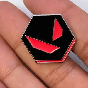 May include: A black hexagonal pin with a red V-shaped logo in the centre.